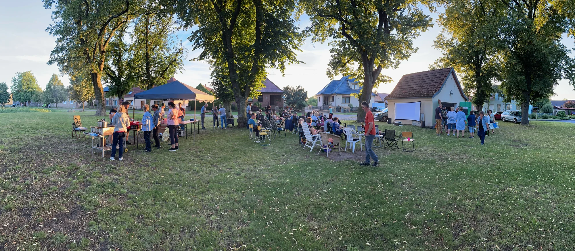Der Dorfanger am Teich beim SommerKino: Campingstühle unter den alten Bäumen, Grillstand mit Pavillon, die Leinwand an der Hauswand.
