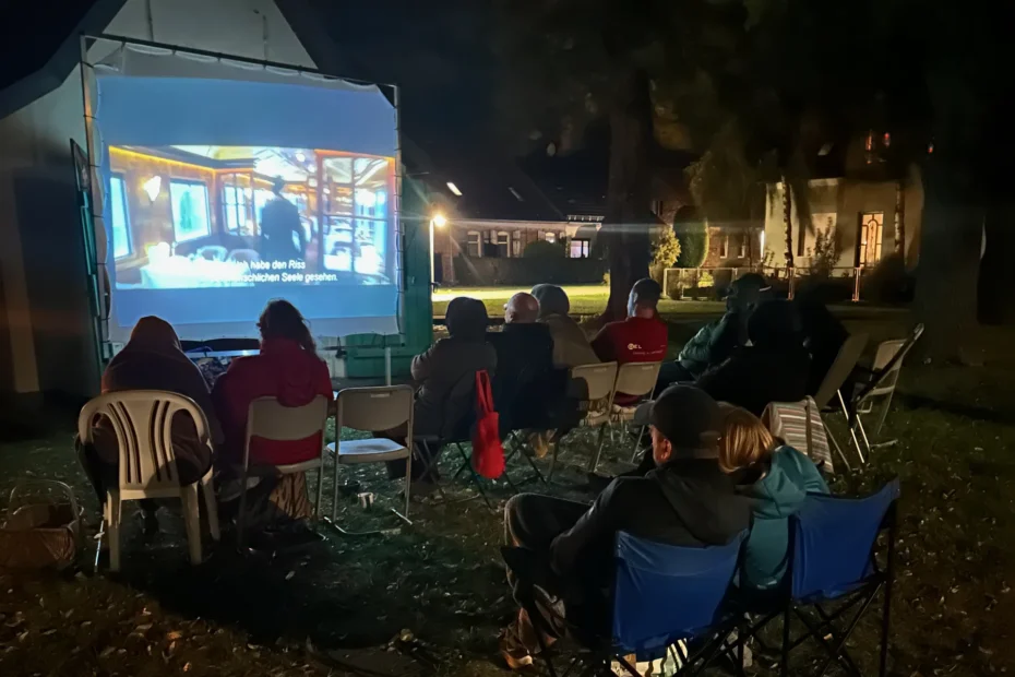 SommerKino in Klosterdorf bei Nacht: Die beleuchtete Leinwand an der Hauswand, davor Zuschauerinnen und Zuschauer in Camping- und Gartenstühlen.